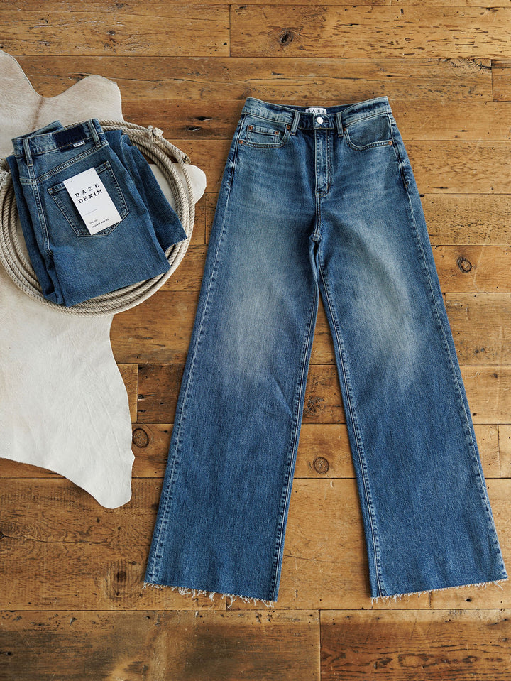The Leyden High Rise Wide Leg Jean by Savannah Sevens western life{&}style, in a 90's-inspired medium wash with frayed hems, is laid flat on a wooden floor beside folded jeans and a rope-filled basket on a white rug.
