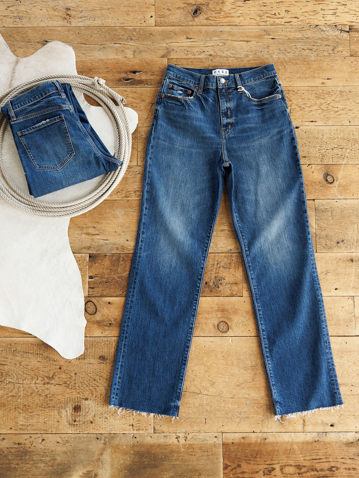 A pair of Daze raw hem jeans and the Savannah Sevens western life{&}style The Payton High Rise Relaxed Straight Leg Jean in medium dark wash denim are laid on a wooden floor with a coiled rope, partially on a white cowhide rug.