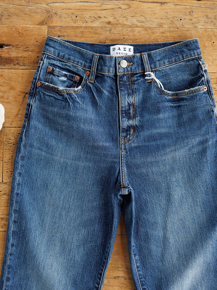 The Payton High Rise Relaxed Straight Leg Jean by Savannah Sevens western life{&}style, in blue, features a button fly, front pockets, and visible stitching, shown laid flat on a wooden surface.