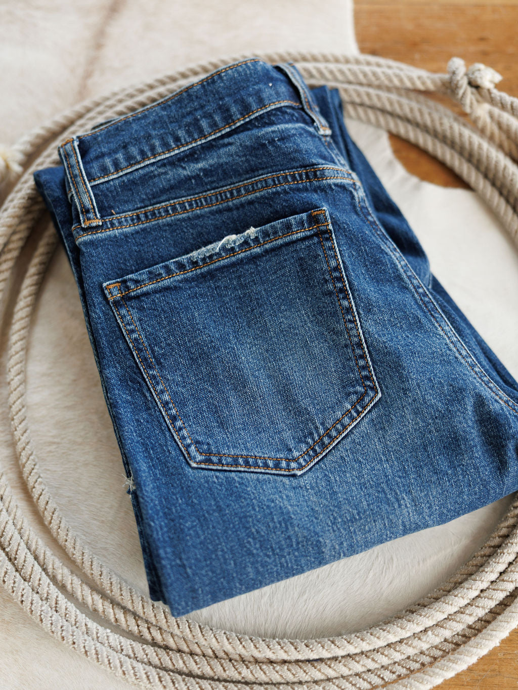 A pair of The Payton High Rise Relaxed Straight Leg Jeans by Savannah Sevens western life{&}style in a medium dark wash is folded on a light-colored animal hide atop a wooden surface.