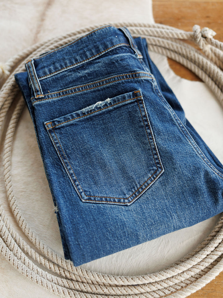 A pair of The Payton High Rise Relaxed Straight Leg Jeans by Savannah Sevens western life{&}style in a medium dark wash is folded on a light-colored animal hide atop a wooden surface.