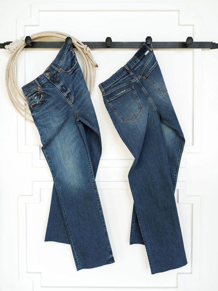 Two pairs of Savannah Sevens western life{&}style The Payton High Rise Relaxed Straight Leg Jeans in medium dark wash hang side by side on a black wall-mounted rack against a white paneled wall, with a coiled rope draped on the left.