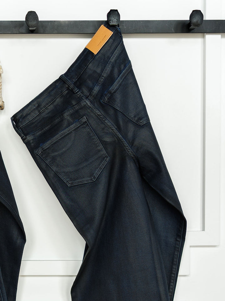 The Riggins High Rise Wide Leg Jean by Savannah Sevens western life{&}style, featuring a coated dark wash and tan leather waistband patch, hangs by a belt loop from a black hook against a white paneled wall.