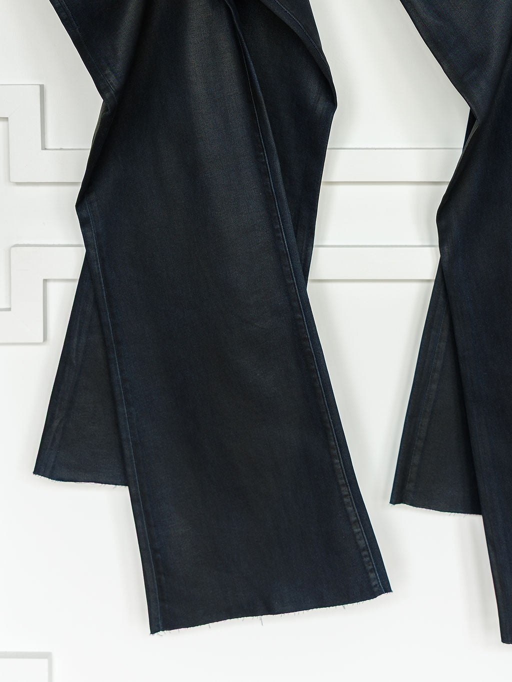 Close-up of The Riggins High Rise Wide Leg Jean by Savannah Sevens western life{&}style, featuring coated dark wash denim, a '90s wide leg silhouette, and raw unfinished hems, hanging against a white textured wall.