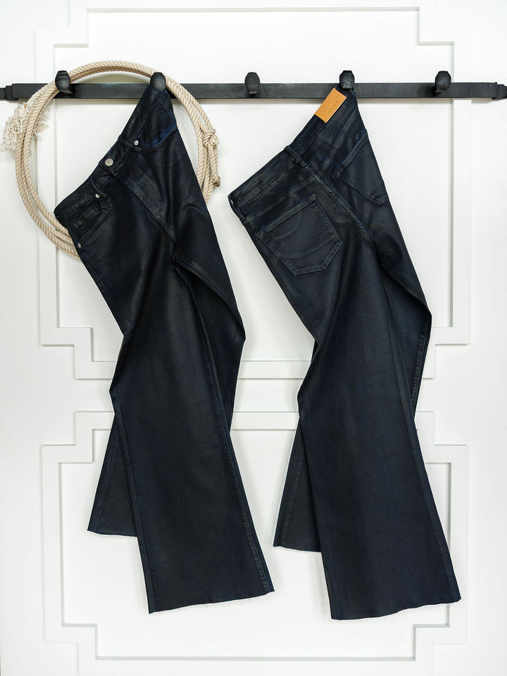 Two pairs of coated dark wash denim jeans, including The Riggins High Rise Wide Leg Jean by Savannah Sevens western life{&}style, hang side by side from a black wall-mounted rack with a light-colored rope on the left, set against a white panel.