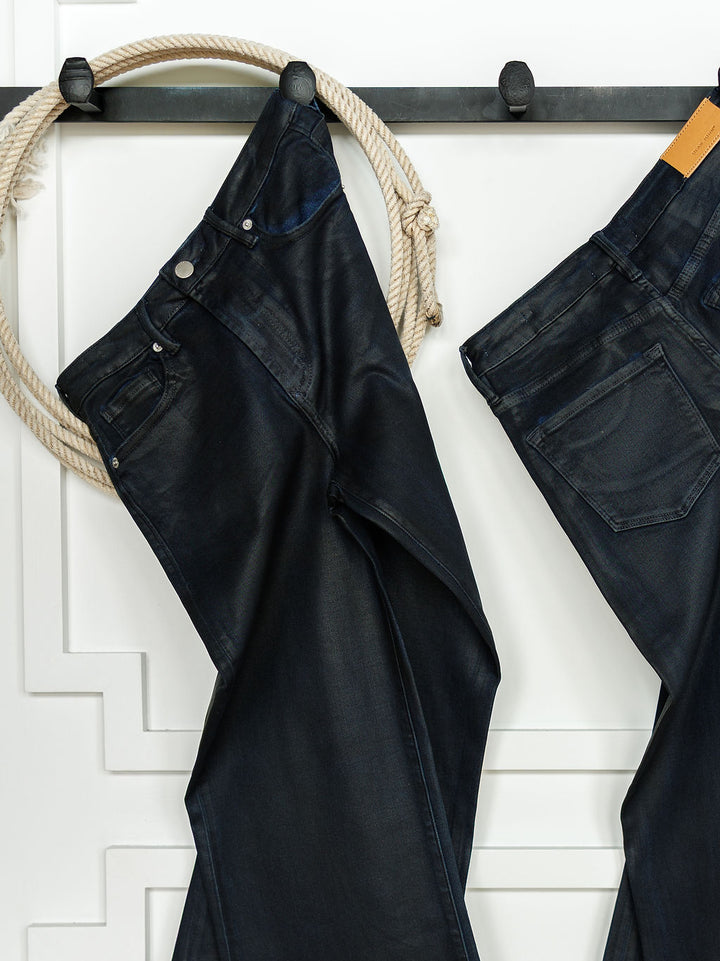 Two pairs of The Riggins High Rise Wide Leg Jean by Savannah Sevens western life{&}style hang from a black wall rack—one draped over a hook with a thick rope, the other by its belt loop—against a white wall with decorative molding.