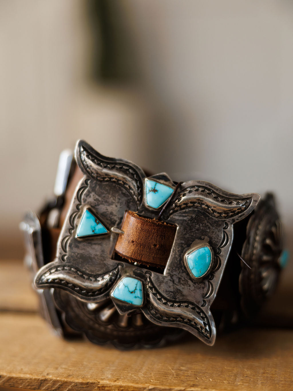 A close-up of the Portales Belt by Savannah Sevens western life{&}style, featuring a vintage sterling silver buckle with engravings and turquoise stones, attached to a Navajo handmade leather belt on a wooden surface.