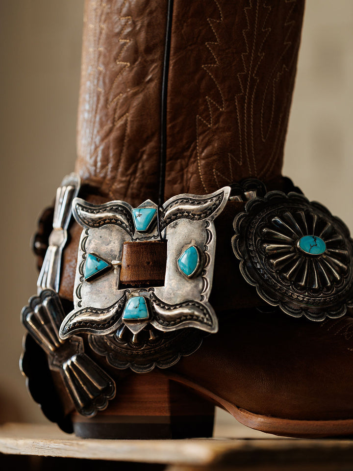 A close-up of the Portales Belt by Savannah Sevens western life{&}style, featuring brown leather with intricate Western detailing, an ornate silver buckle, and turquoise concho accents.