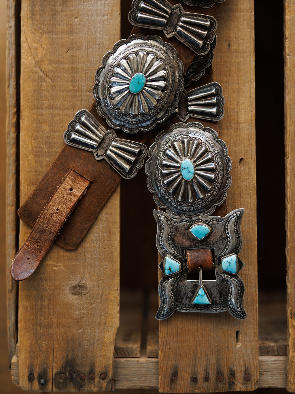 The Portales Belt by Savannah Sevens western life{&}style features brown leather with large, ornate silver conchos and a rectangular buckle adorned with turquoise stones, capturing vintage Southwestern style on a rustic wooden surface.