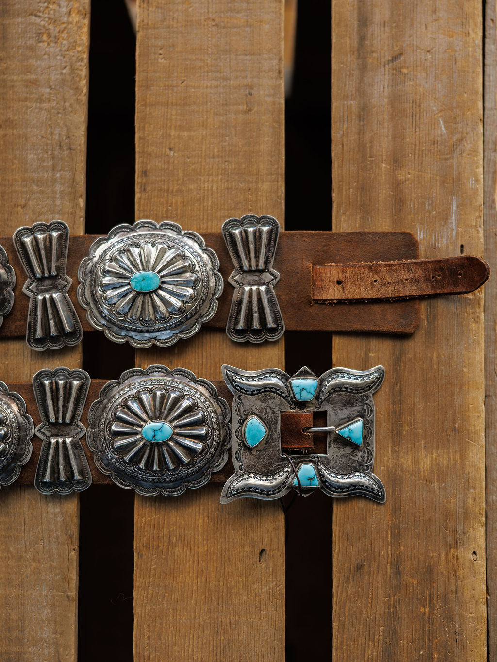 The Portales Belt by Savannah Sevens western life{&}style features large, ornate sterling silver conchos with #8 Turquoise stones and a rectangular buckle, shown on a wooden surface.