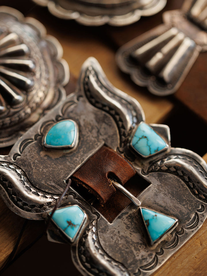 A close-up of the Portales Belt by Savannah Sevens western life{&}style, featuring a vintage sterling silver buckle with turquoise stones on brown leather, shown with other detailed silver accessories in the background.