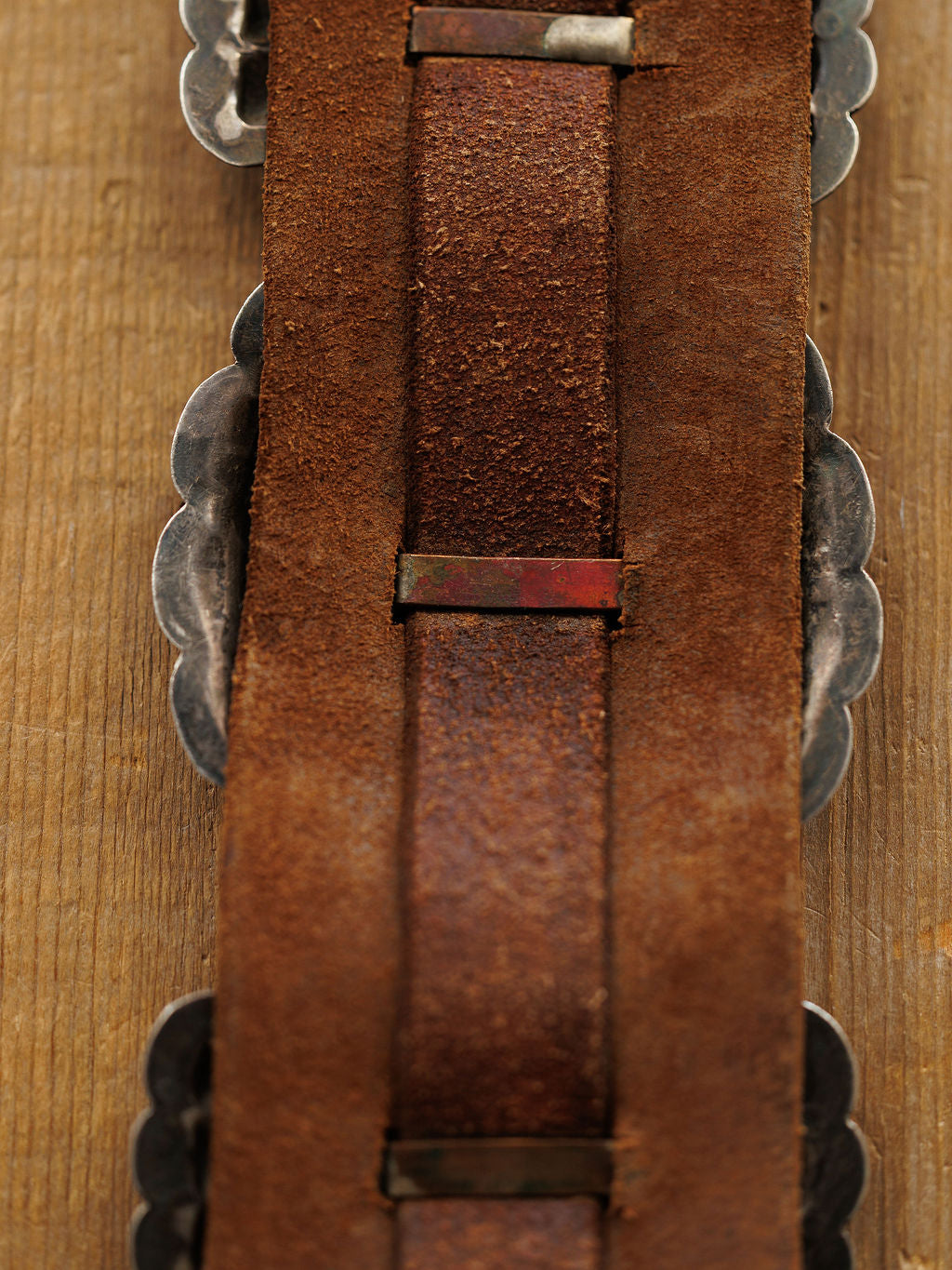 Close-up of the back side of the Portales Belt by Savannah Sevens western life{&}style, featuring worn brown leather with metal accents and scalloped edges, displayed on a wooden surface.