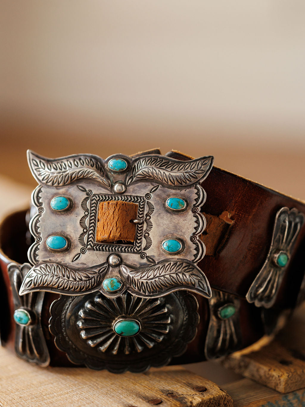 A close-up of the New Mexico Belt by Savannah Sevens western life{&}style, showcasing handmade leather with ornate silver and turquoise conchos, detailed engravings, and leaf motifs on a wooden surface.