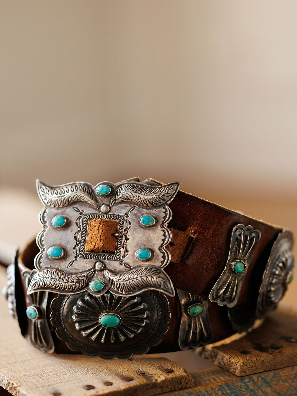 The New Mexico Belt by Savannah Sevens western life{&}style features ornate silver conchos and a large turquoise-accented buckle, beautifully displayed on a rustic wooden surface.