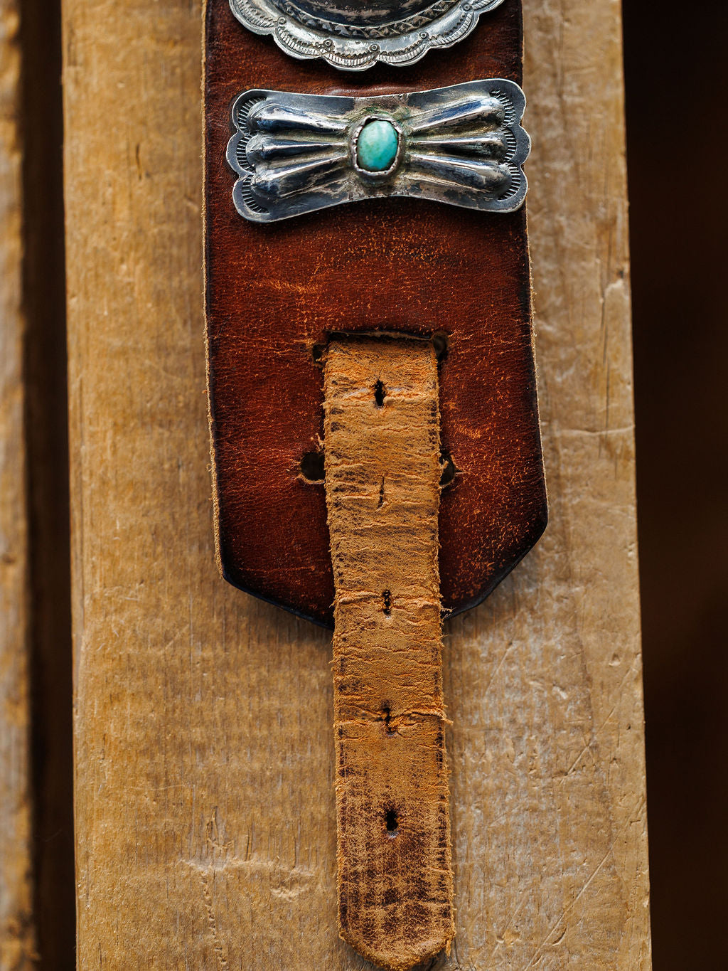 A close-up of the New Mexico Belt by Savannah Sevens western life{&}style features a sterling silver turquoise buckle, shown against wood. The handmade leather displays creases and holes from frequent use.