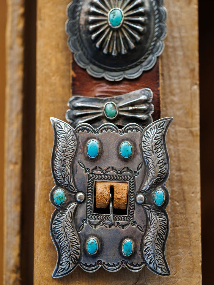 A close-up of the New Mexico Belt by Savannah Sevens western life{&}style, featuring a vintage-inspired sterling silver turquoise buckle on a handmade leather concho belt, displayed against a wooden background.
