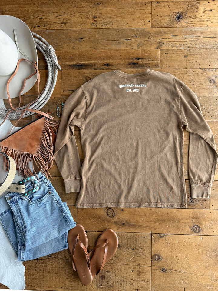Flat lay of the S7s & Co. Exclusive Graphic Top by Savannah Sevens western life{&}style—a tan, relaxed fit, long sleeve mineral wash shirt featuring “SAVANNAH SEVENS EST. 2013”—styled with a hat, sunglasses, fringe bag, denim shorts, beaded belt, jewelry, and brown sandals on a wooden floor.