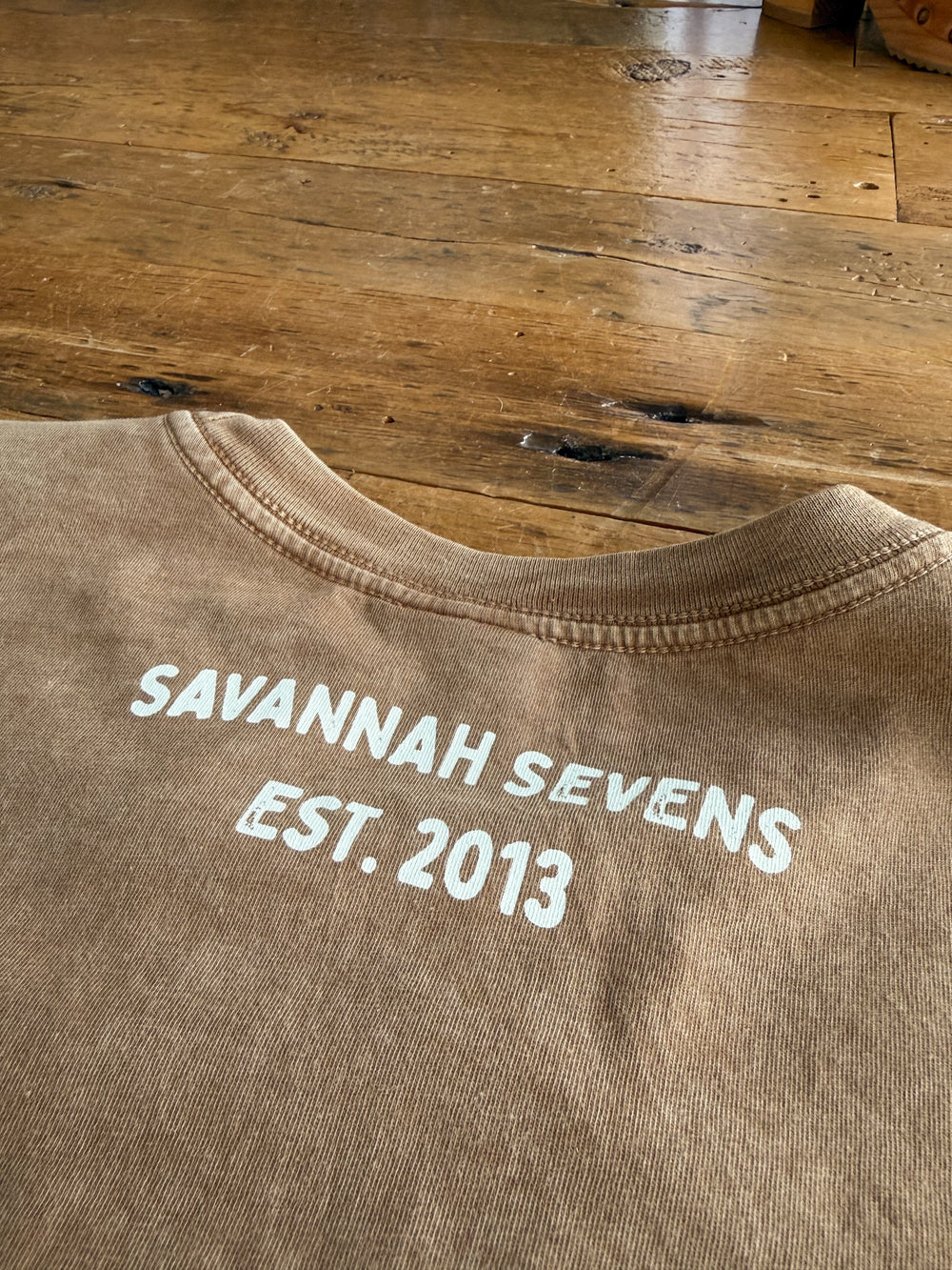 A S7s & Co. Exclusive Graphic Top from Savannah Sevens western life{&}style rests on a wooden floor, featuring white “SAVANNAH SEVENS EST. 2013” text printed on the back near the collar. This women’s relaxed fit shirt combines comfort with standout western-inspired style.