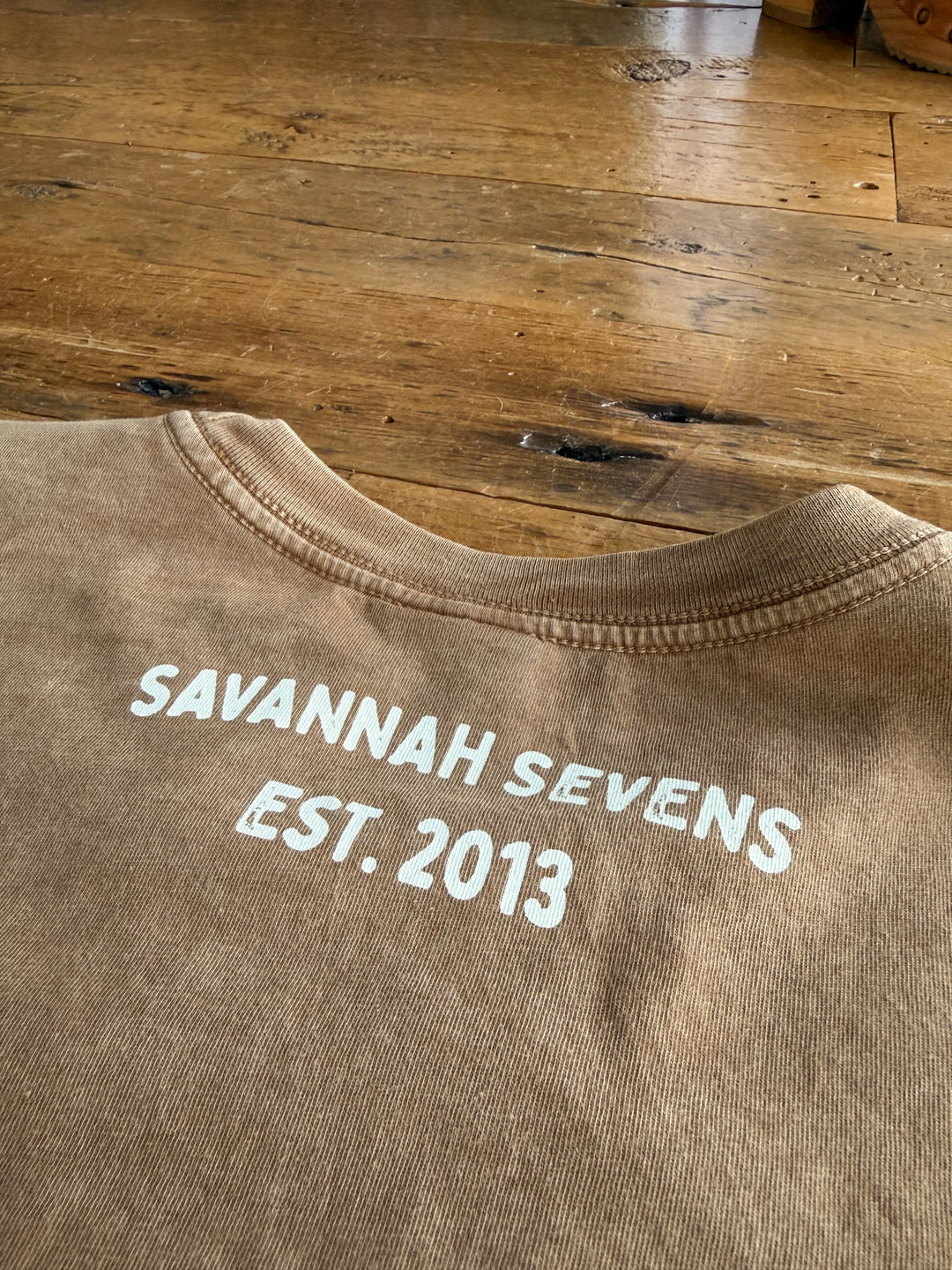 A S7s & Co. Exclusive Graphic Top from Savannah Sevens western life{&}style rests on a wooden floor, featuring white “SAVANNAH SEVENS EST. 2013” text printed on the back near the collar. This women’s relaxed fit shirt combines comfort with standout western-inspired style.
