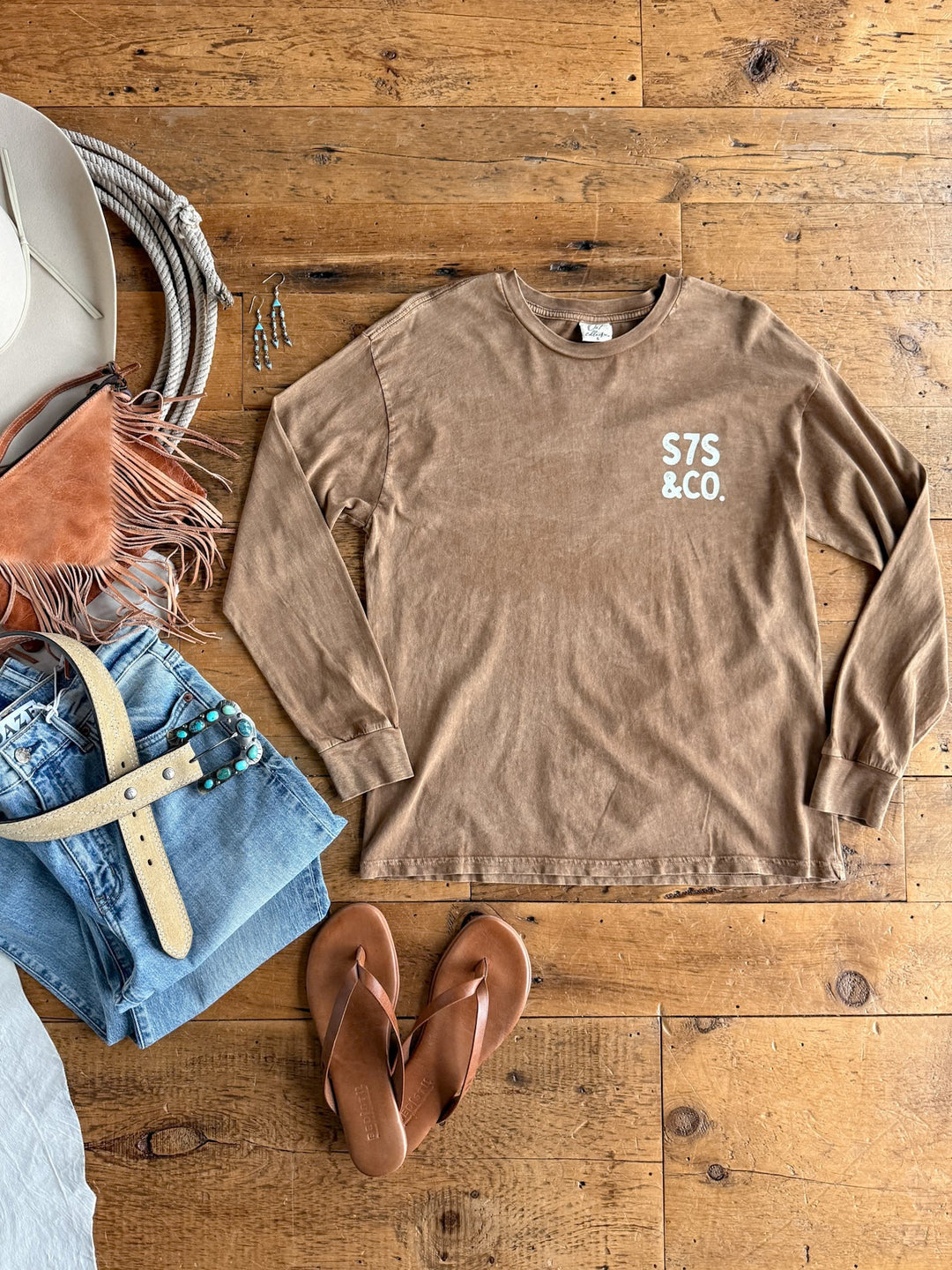 Flat lay of a brown S7s & Co. Exclusive Graphic Top from Savannah Sevens western life{&}style, paired with blue jeans featuring a beige belt, sandals, a fringed bag, hat, and jewelry, all arranged on a wooden floor.