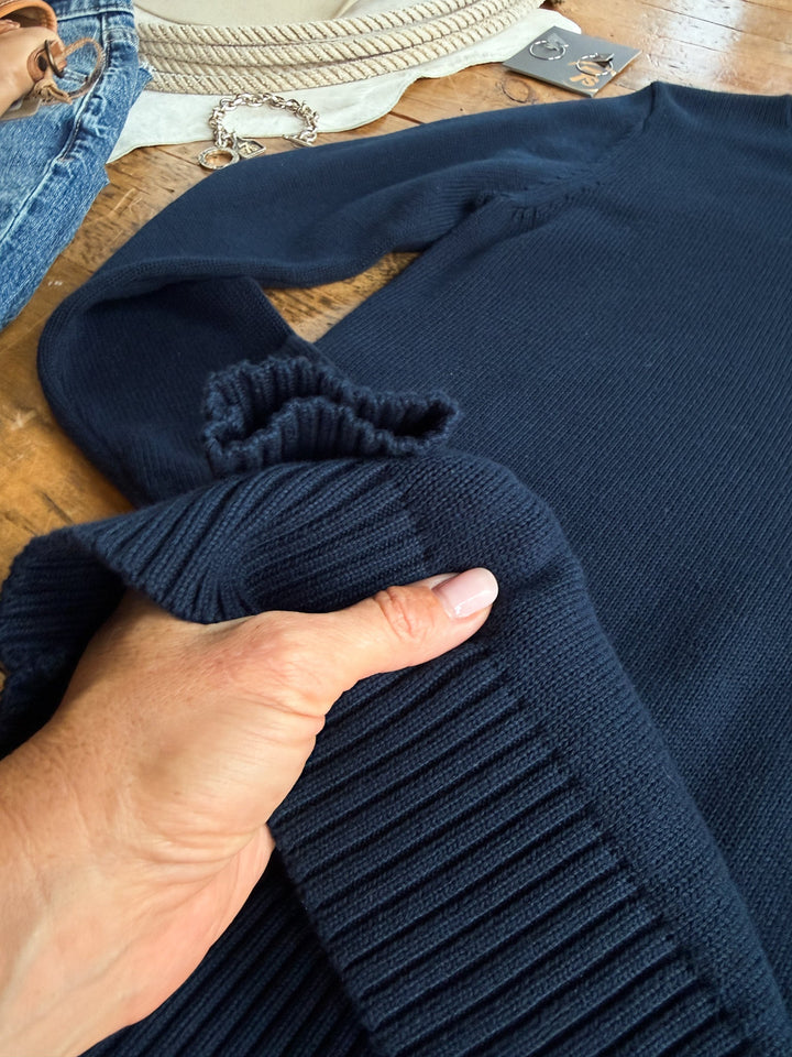 A hand with pale pink nails holds the ribbed cuff of the S7s & Co. Navy Varsity Sweater by Savannah Sevens western life{&}style on a wooden table, surrounded by jeans, jewelry, and other accessories.