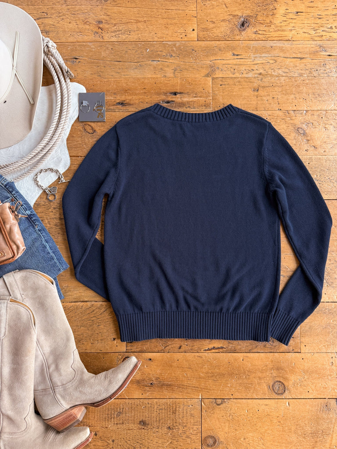 A S7s & Co. Navy Varsity Sweater from Savannah Sevens western life{&}style lies flat on a wooden floor beside beige cowboy boots, a white hat, coiled rope, denim shorts, a bracelet, and a black tag. This exclusive cotton sweater is available only at Savannah Sevens.