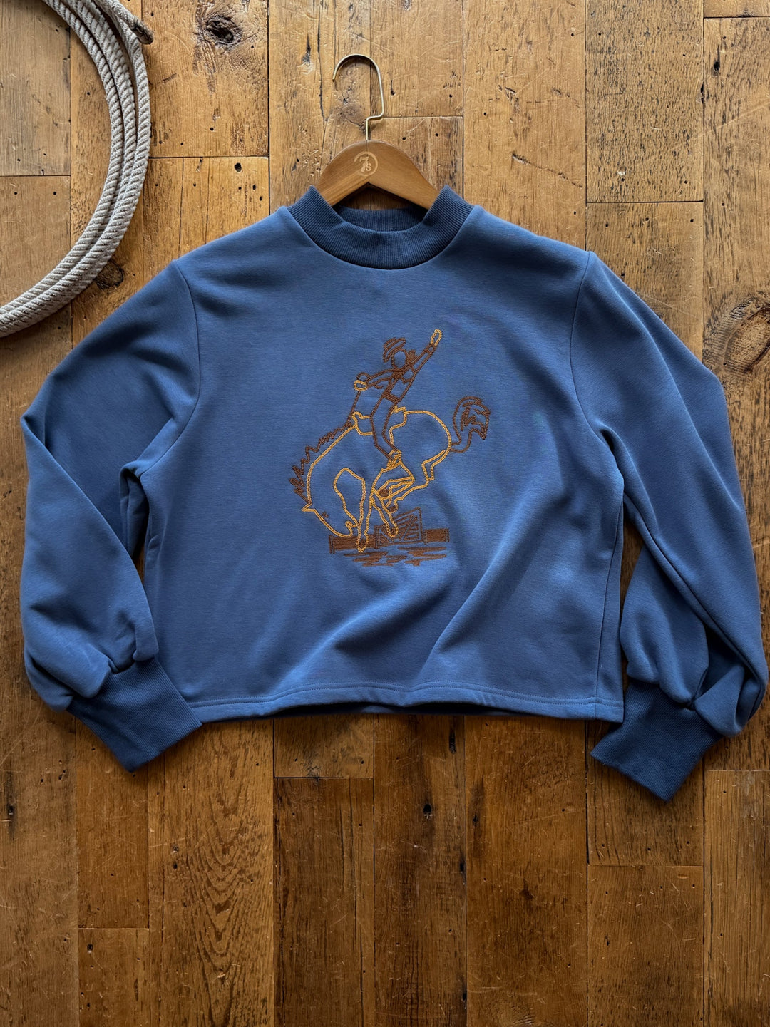 Savannah Sevens western life&style Saddle Bronc Slate Blue Embroidered Pullover, a slate blue fleece-lined mock-neck featuring a gold chain-stitch embroidered graphic of a cowboy riding a bucking bronc.