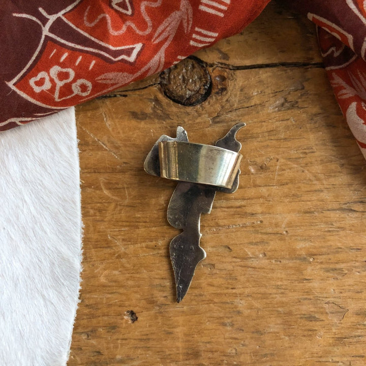 The Saddle Scarf Slide by Savannah Sevens western life{&}style, featuring a jagged abstract design in German silver, is displayed on a wooden surface beside white fur and red patterned fabric.