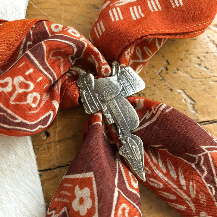 A red and brown patterned bandana is secured with the Saddle Scarf Slide by Savannah Sevens western life{&}style, a German silver accessory engraved with western motifs, resting on a wooden surface.