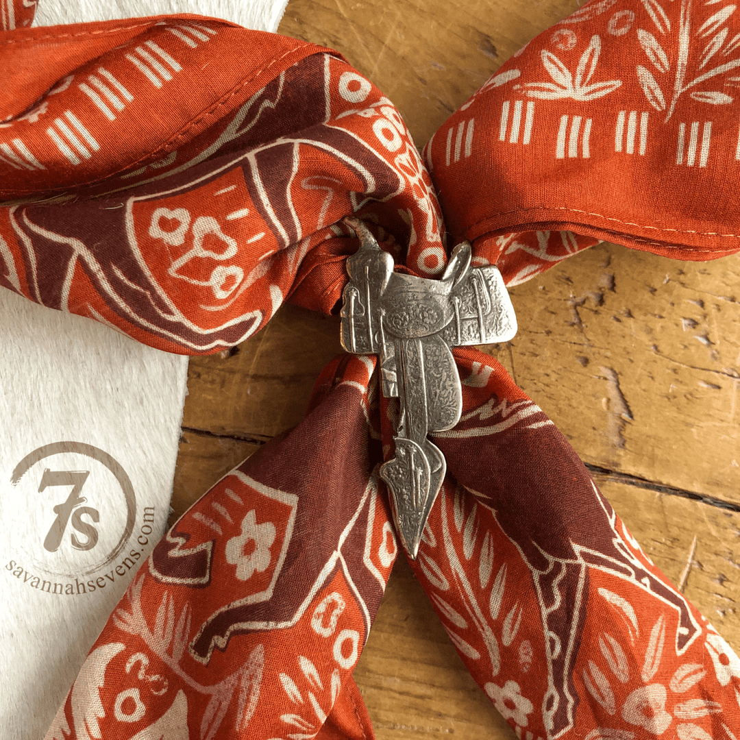 A red patterned bandana with the Saddle Scarf Slide from Savannah Sevens western life{&}style rests on wood beside a white cloth displaying the savannahsevens.com logo and 7s.