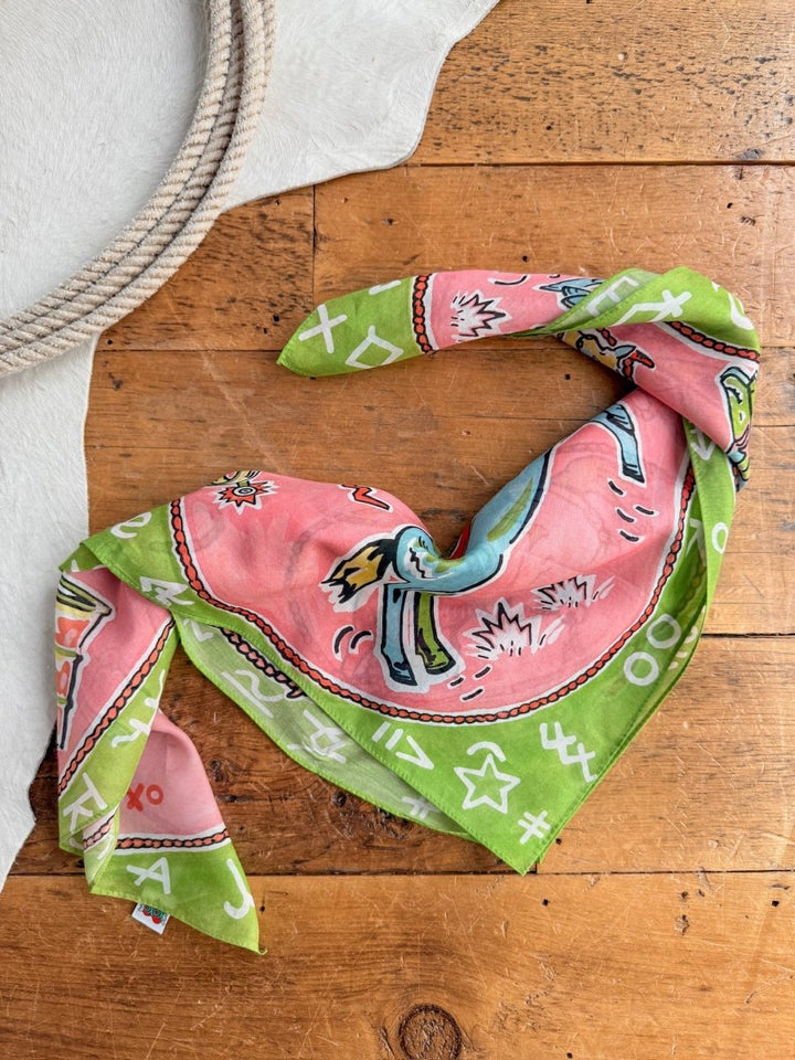 The Saddle Up Neck Scarf by Savannah Sevens western life{&}style—featuring green and pink retro western designs—rests on a wooden surface beside a rope and white cowhide rug.