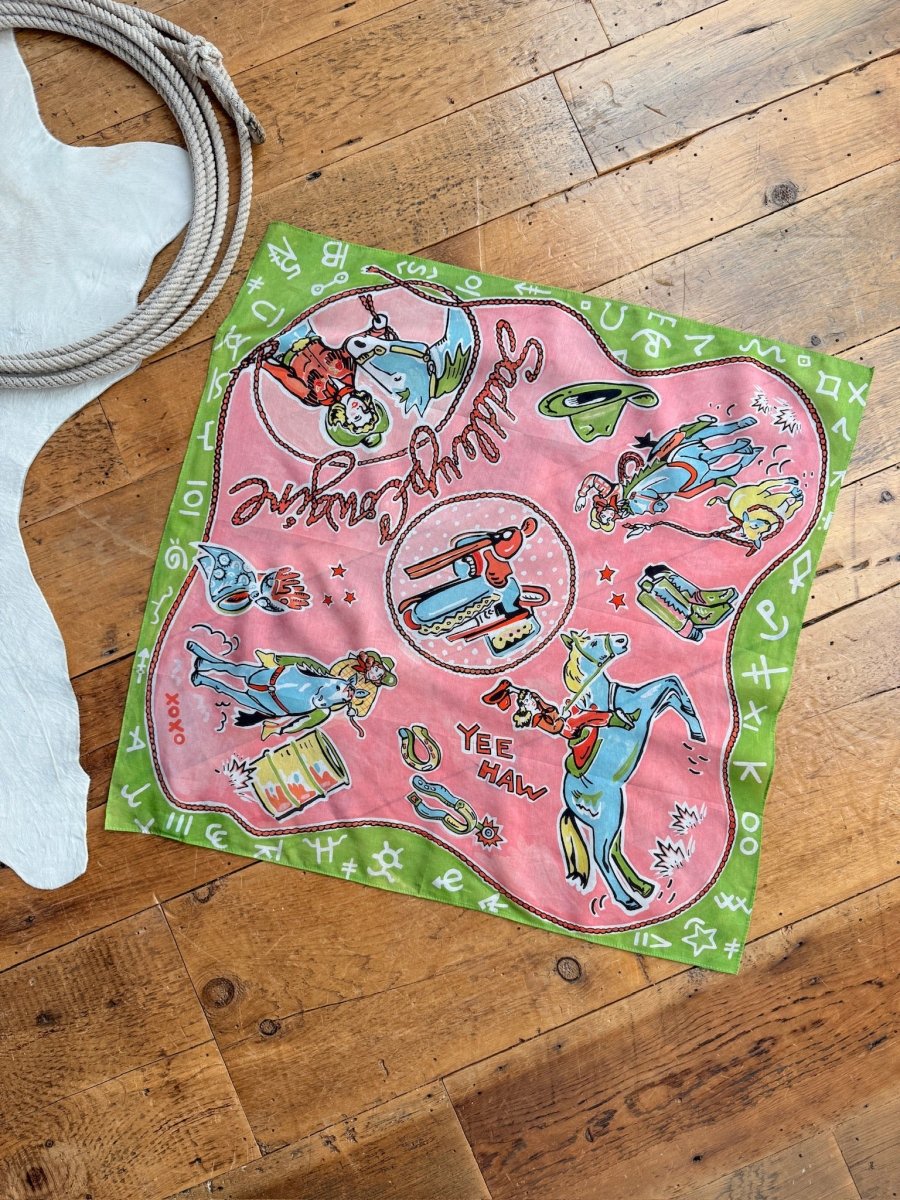 The Saddle Up Neck Scarf by Savannah Sevens western life{&}style features colorful cowboy illustrations, boots, horses, and “Saddle up, partner! YEE HAW.” The cotton-silk scarf is displayed on a wooden floor with rope and a white fur rug.