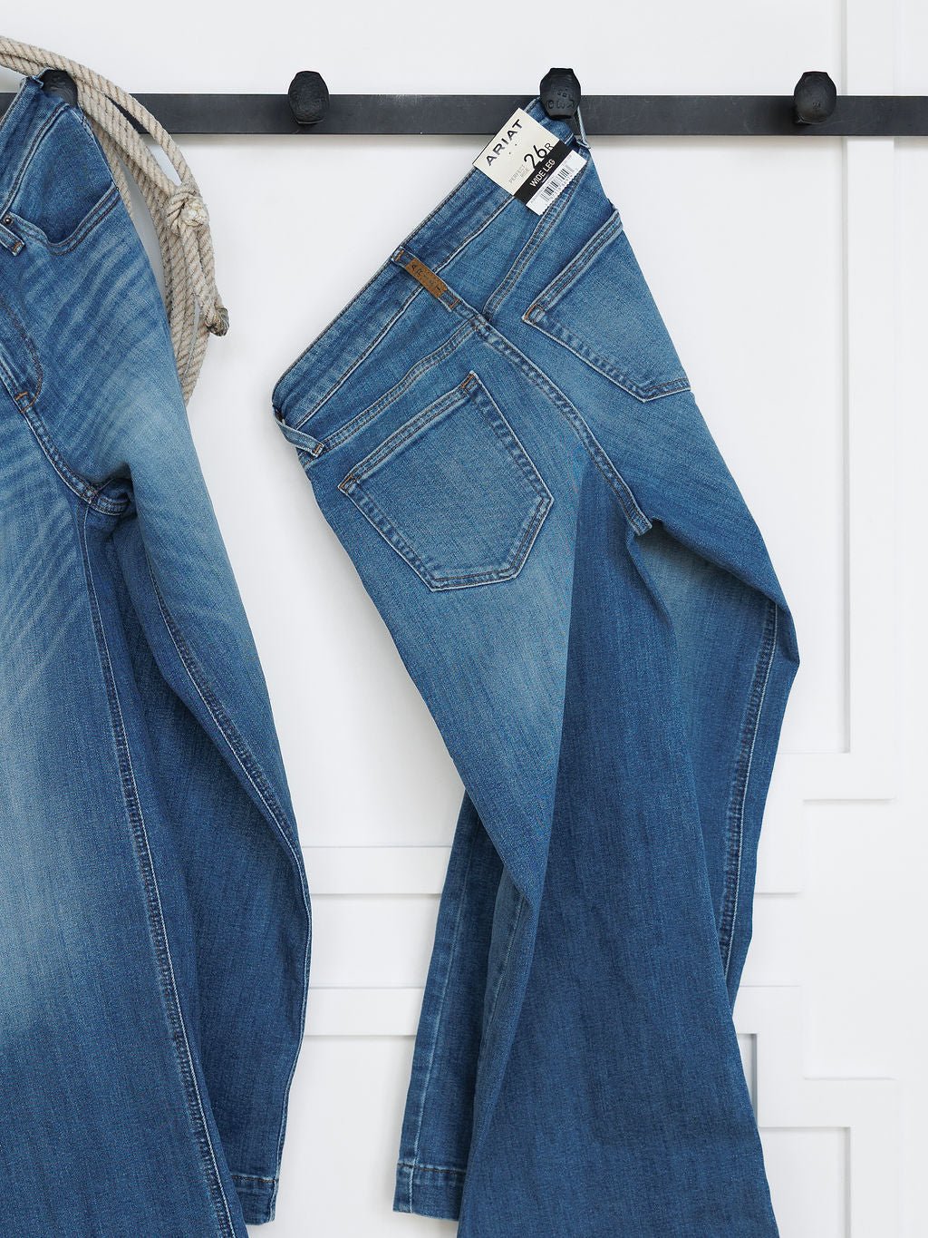 The Sadie Perfect Rise Wide Leg Crop Jean by Savannah Sevens western life{&}style in a medium light wash hangs on a black wall hook beside another pair of jeans and a striped rope, set against a white wall with decorative molding.