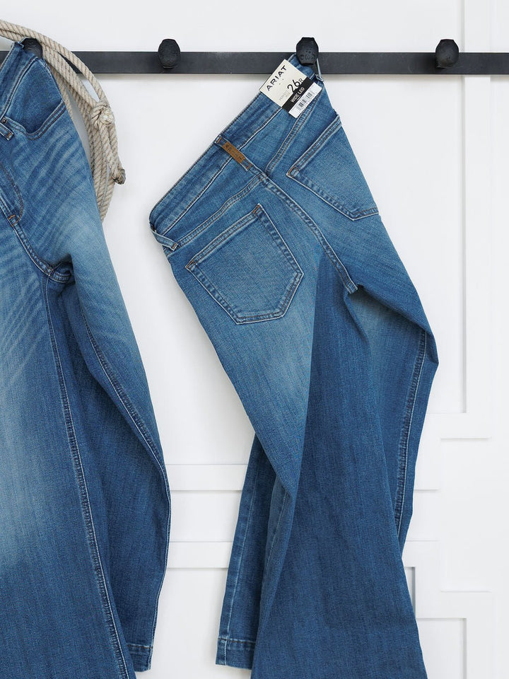 The Sadie Perfect Rise Wide Leg Crop Jean by Savannah Sevens western life{&}style in a medium light wash hangs on a black wall hook beside another pair of jeans and a striped rope, set against a white wall with decorative molding.