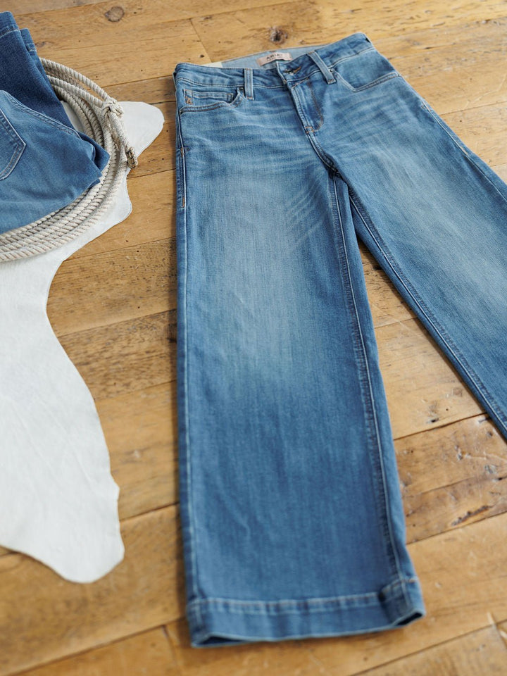A pair of Savannah Sevens western life{&}style The Sadie Perfect Rise Wide Leg Crop Jeans in a medium light wash is laid flat on a wooden floor beside a white animal hide and a coiled rope.