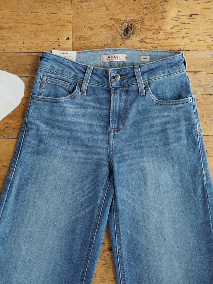 The Sadie Perfect Rise Wide Leg Crop Jean by Savannah Sevens western life{&}style in a medium light wash is laid flat on a wooden surface, showcasing the perfect rise waistband, front pockets, fly, and belt loops.