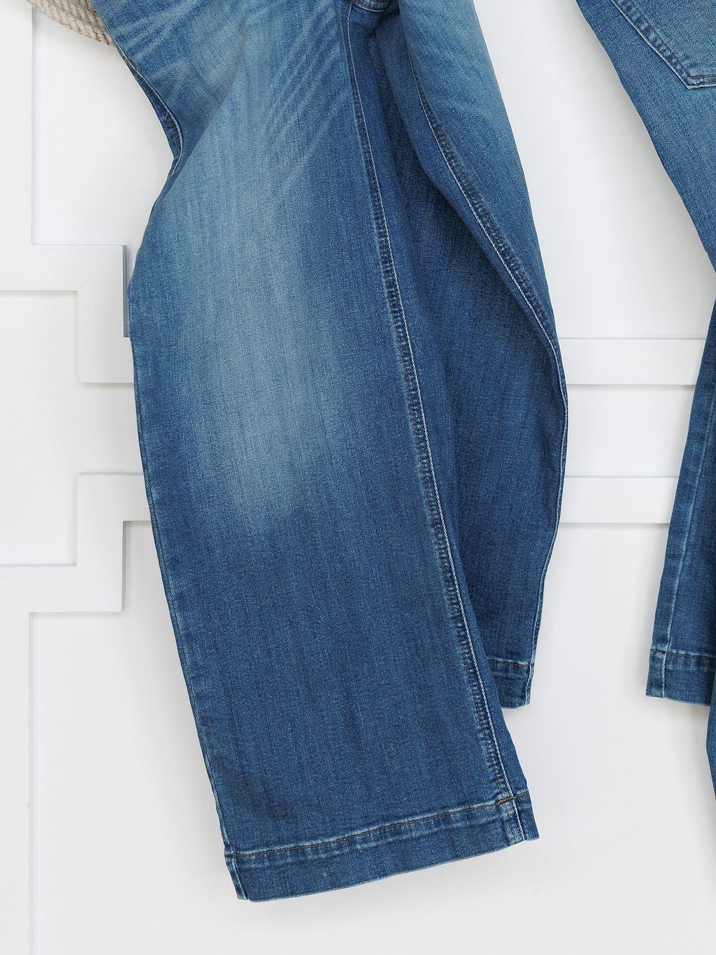 A close-up of the lower legs of The Sadie Perfect Rise Wide Leg Crop Jean by Savannah Sevens western life{&}style in a medium light wash, featuring a faded effect and visible seam stitching, hanging against a white paneled wall.