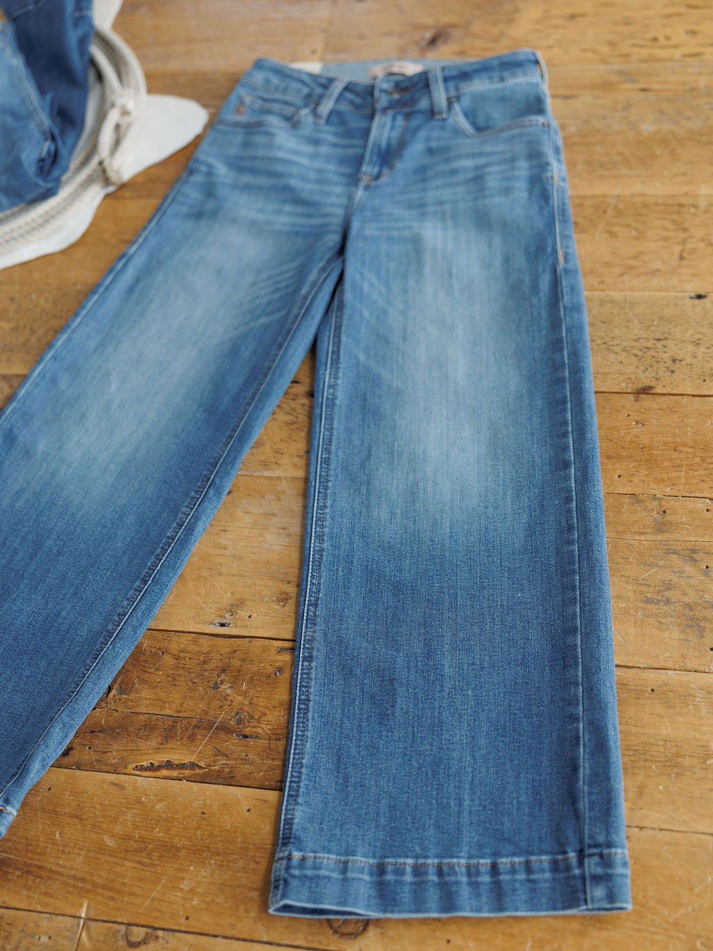 The Sadie Perfect Rise Wide Leg Crop Jean by Savannah Sevens western life{&}style, in a medium light wash, is laid flat on a wooden floor with a glimpse of a denim jacket visible in the corner.