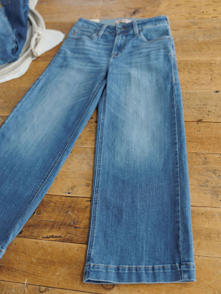 The Sadie Perfect Rise Wide Leg Crop Jean by Savannah Sevens western life{&}style, in a medium light wash, is laid flat on a wooden floor with a glimpse of a denim jacket visible in the corner.