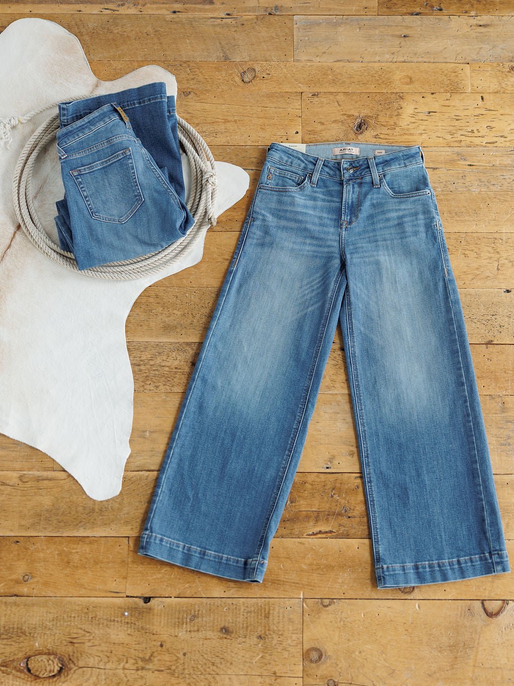 Two pairs of blue jeans on wood: one folded with a rope atop a cowhide rug, the other flat to show wide legs and a light wash. Explore The Sadie Perfect Rise Wide Leg Crop Jean by Savannah Sevens western life{&}style for effortless style.