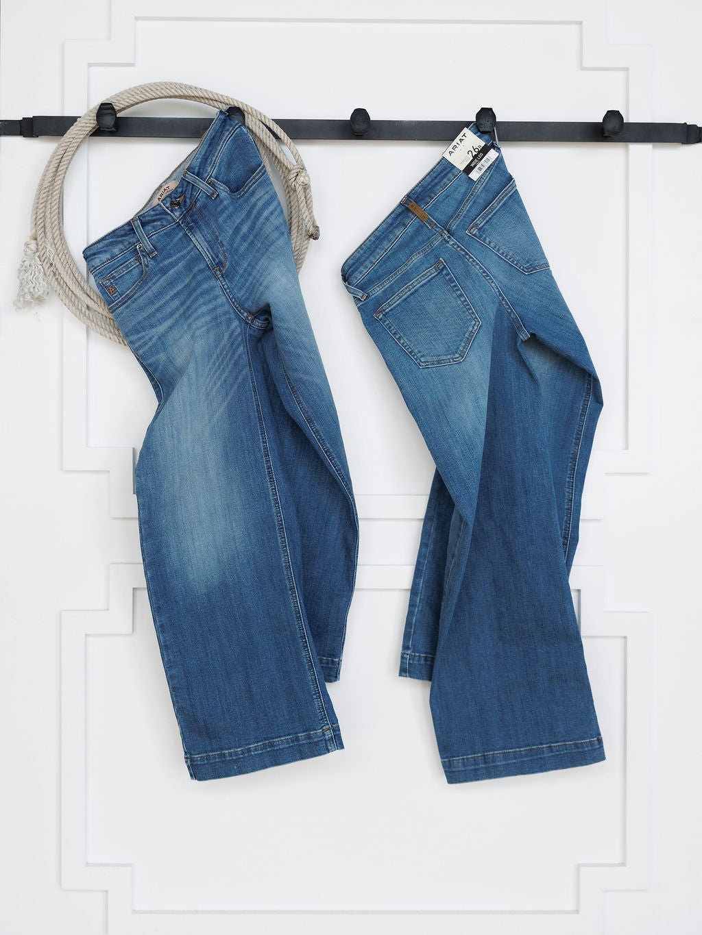 Two pairs of Savannah Sevens western life{&}style The Sadie Perfect Rise Wide Leg Crop Jeans in medium light wash hang on a black wall rack with a coiled rope, against a white panel. Each pair features slight differences in rise and fit.