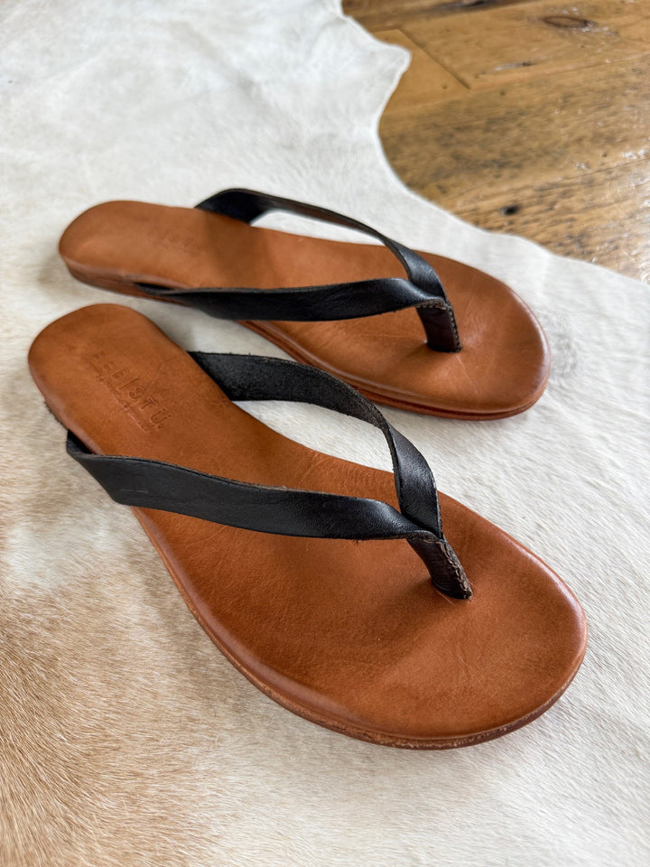 A pair of Sadler Black Leather Flip Flop sandals by Savannah Sevens western life{&}style, featuring black leather straps and a cushioned leather footbed, are placed on a light-colored cowhide rug atop a wooden floor.