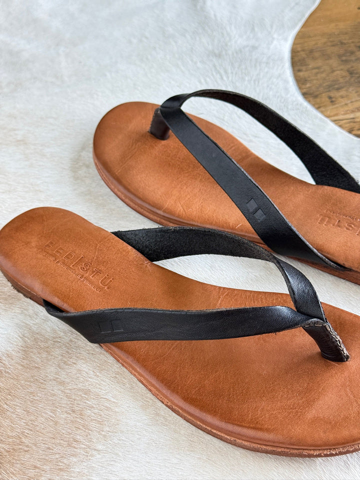 A pair of Sadler Black Leather Flip Flop sandals by Savannah Sevens western life{&}style, featuring a cushioned leather footbed and black leather straps, resting on a light-colored textured surface.