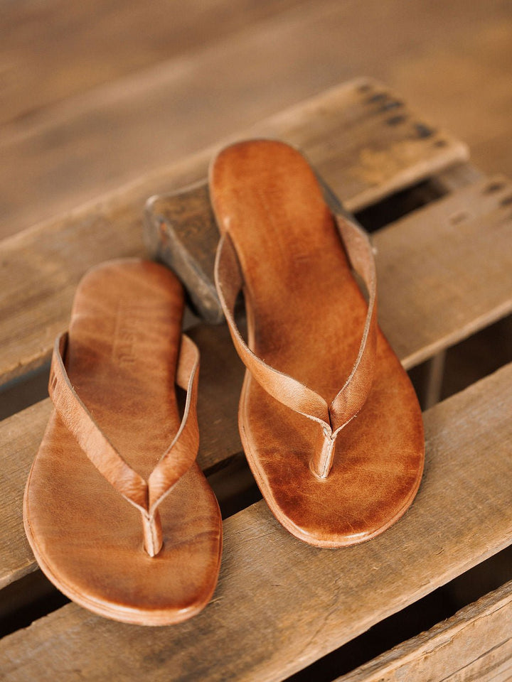 The Sadler leather flip flop sandals by Savannah Sevens western life{&}style, featuring a cushioned footbed, are displayed on a rustic wooden surface. Sandals made by BedStu. 