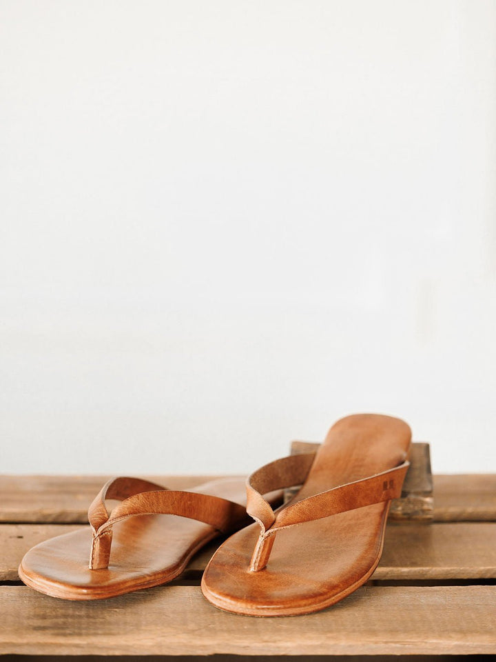A pair of The Sadler leather flip flops by Savannah Sevens western life{&}style, featuring a cushioned footbed, rests on a wooden surface against a plain, light background. Sandals by BedStu.