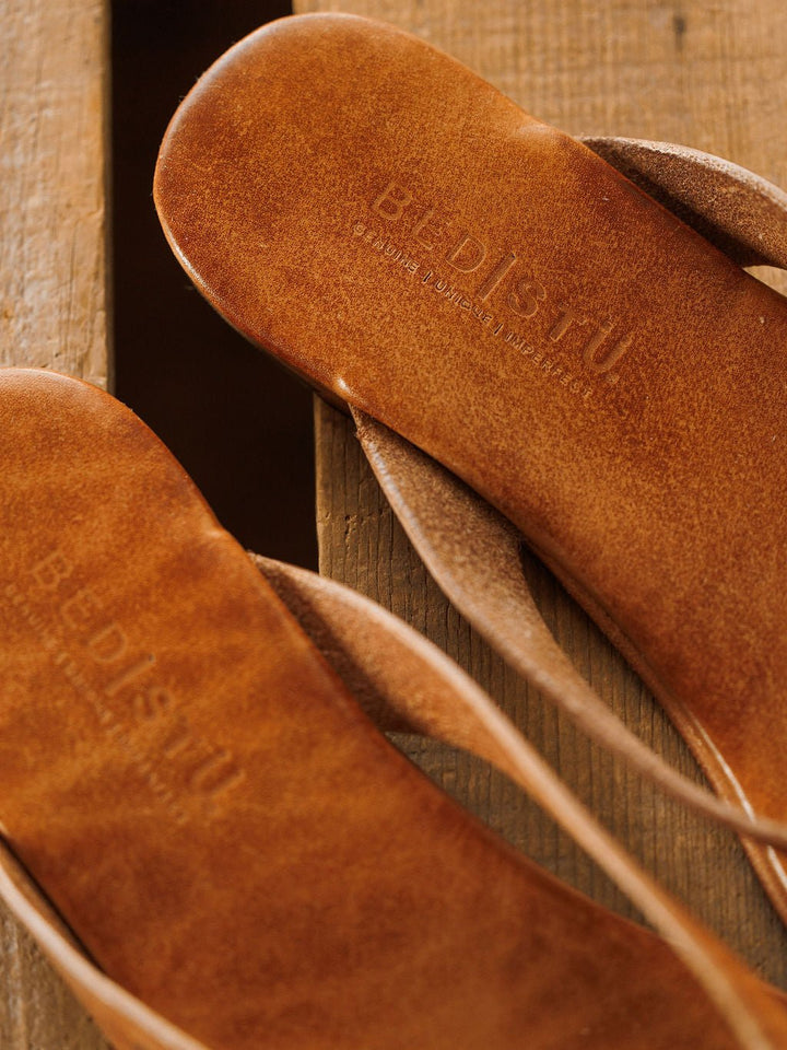Close-up of two brown The Sadler flip flops by Savannah Sevens western life{&}style on a wooden surface, with the brand embossed on the insole. The texture, craftsmanship, and cushioned footbed are clearly visible. Sandals by BedStu.