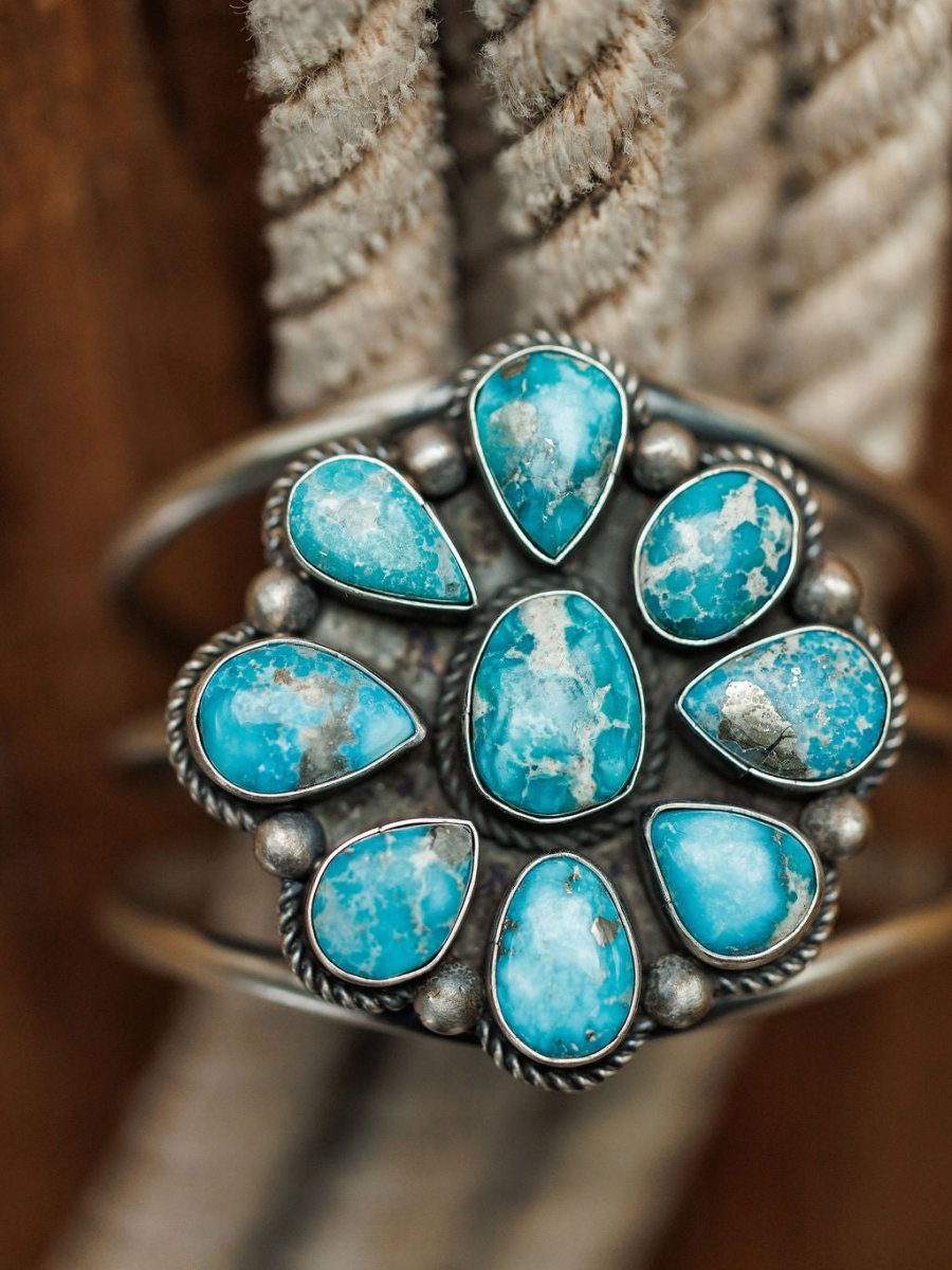 The Sagola Cuff by Savannah Sevens western life{&}style is a unique sterling silver cuff with a floral design and nine turquoise stones in a circular pattern, displayed on coiled beige rope.