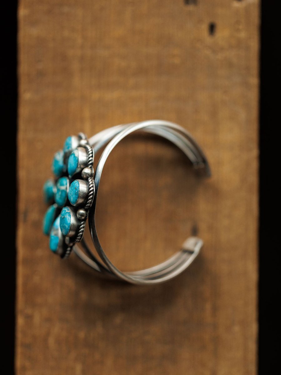 The Sagola Cuff by Savannah Sevens western life{&}style is a unique sterling silver bracelet with a cluster of blue turquoise stones, elegantly showcased on a rustic wooden surface.
