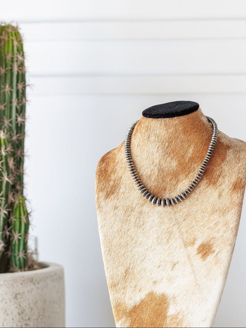 The Saledo Necklace by Savannah Sevens western life{&}style, a 20.5-inch sterling silver Navajo pearl necklace with graduated rondelle beads, is displayed on a cowhide-patterned jewelry bust with a potted cactus and white wall in the background.