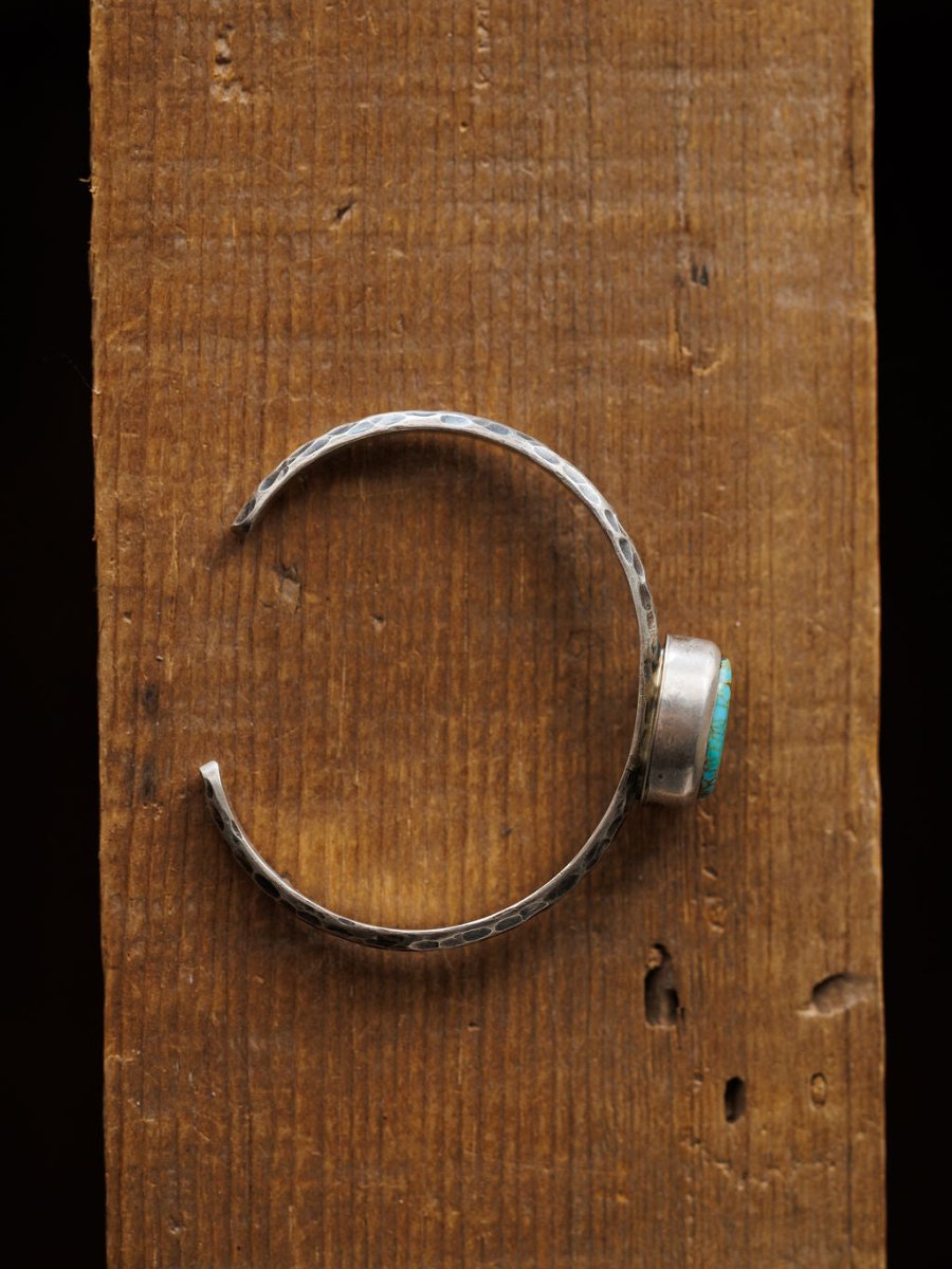 The Salida Cuff by Savannah Sevens western life{&}style features a hammered open-end design and a round Kings Manassa turquoise stone, showcased on a rustic wooden surface.